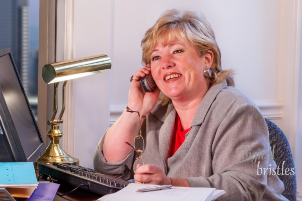 Mature businesswoman talking on the phone with reading glasses in hand and piles of paperwork