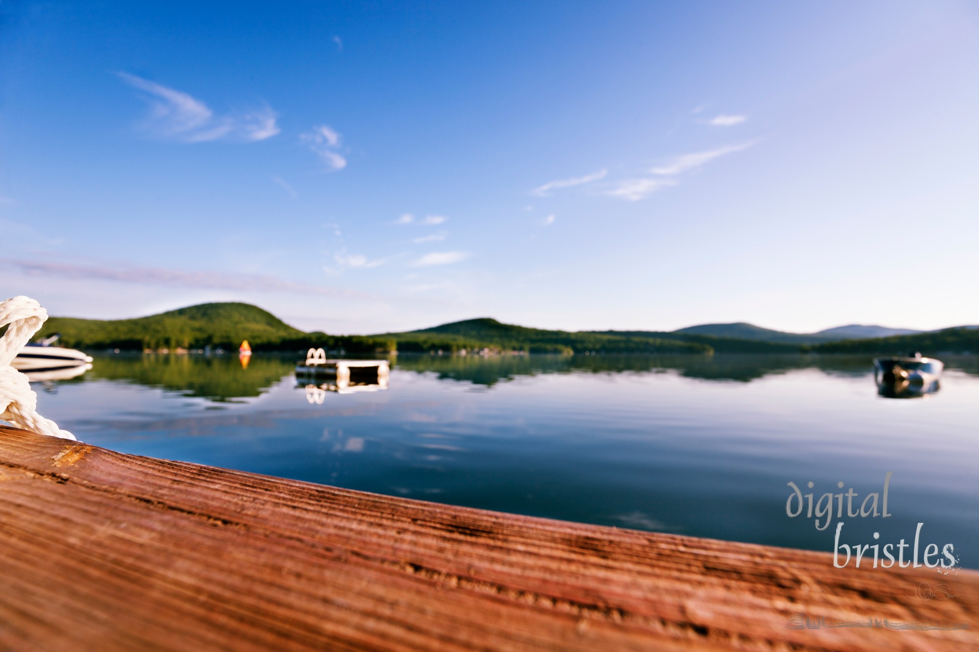 View of a peaceful lake from the end of a dock. Focus on the rope & edge of the dock